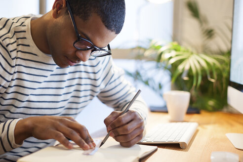 Junger kreativer Mann sitzt am Schreibtisch und schreibt in sein Notizbuch - EBSF000542