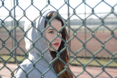 Portrait of woman with hooded jacket behind mesh wire fence - BFRF001065