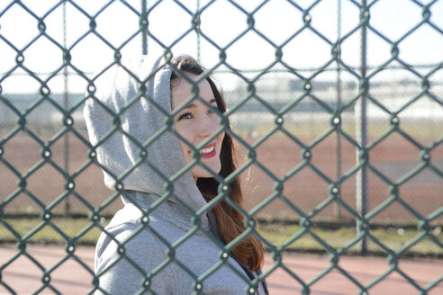 Portrait of woman wearing hooded jacket behind mesh wire fence - BFRF001066