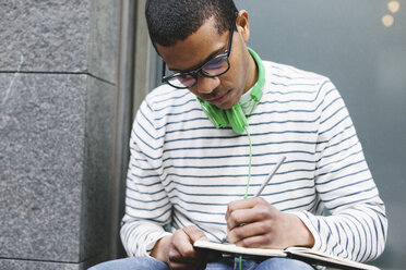 Junger Mann mit grünen Kopfhörern sitzt draußen und schreibt etwas in sein Notizbuch - EBSF000561
