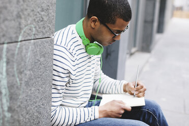 Junger Mann mit grünen Kopfhörern sitzt draußen und schreibt etwas in sein Notizbuch - EBSF000562