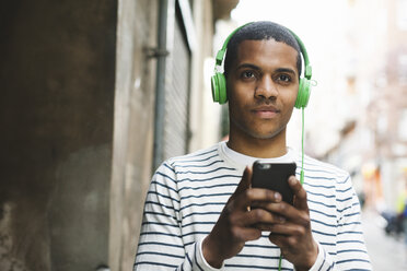 Spanien, Barcelona, Porträt eines lächelnden jungen Mannes, der mit grünen Kopfhörern auf der Straße Musik hört - EBSF000574