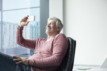 Man sitting in a leather chair taking a selfie with his smartphone - RBF002623