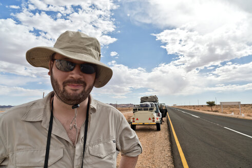 Namibia, Mann mit Safari-Hut und Sonnenbrille mit Auto am Straßenrand im Hintergrund - CLPF000105