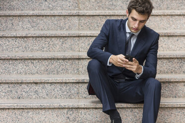 Porträt eines jungen Geschäftsmannes, der auf einer Treppe sitzt und ein Smartphone benutzt - ABZF000004