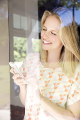 Blonde Frau mit Smartphone am Fenster stehend - TOYF000100