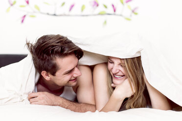 Laughing couple lying side by side in bed under blanket - DAWF000346