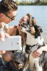 Glückliches junges Paar macht Selfie mit Hund am Flussufer - UUF003938