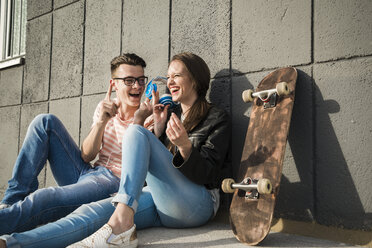 Happy young couple sitting on ground with headphones and skateboard - UUF003898