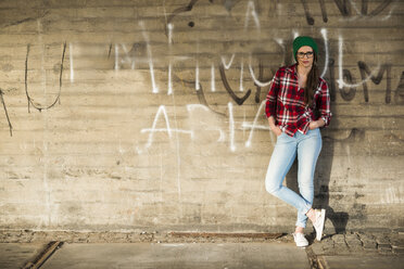 Young woman with checkered shirt and wooly hat leaning at graffiti wall - UUF003909