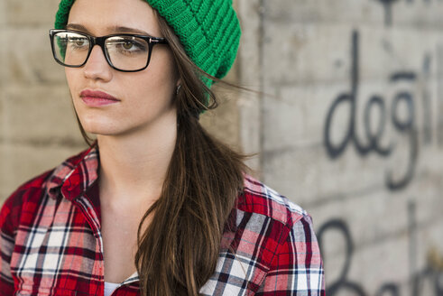 Young woman with checkered shirt and green wooly hat at graffiti wall - UUF003910