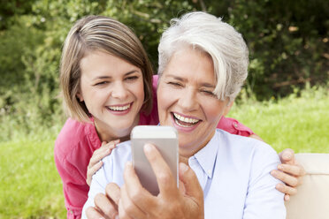 Lachende Mutter mit erwachsener Tochter, die im Freien auf ihr Handy schaut - MFRF000160