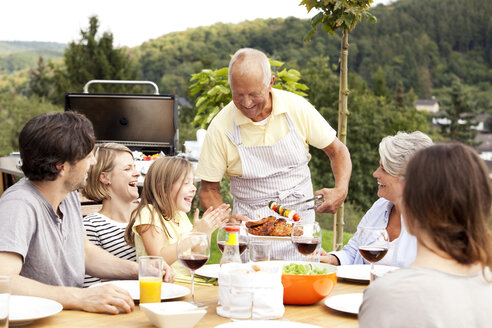 Grandfather serving food from barbecue grill for family at garden table - MFRF000203