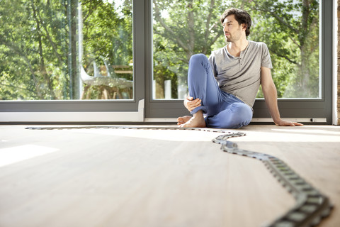 Nachdenklicher Mann auf dem Boden sitzend mit den Schienen einer Spielzeugeisenbahn, lizenzfreies Stockfoto