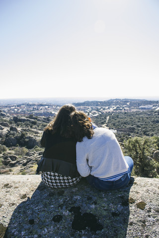 Spanien, Torrelodones, zwei Freundinnen sitzen nebeneinander auf einem Felsen und betrachten die Aussicht, lizenzfreies Stockfoto