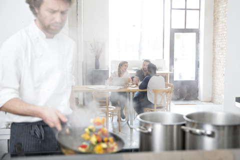 Koch bei der Arbeit in der Küche seines Restaurants, während die Gäste im Hintergrund kommunizieren, lizenzfreies Stockfoto
