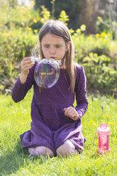 Mädchen hockt auf einer Wiese und bläst eine große Seifenblase - SARF001754
