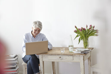 Reife Frau sitzt am Tisch und benutzt einen Laptop - FMKF001462