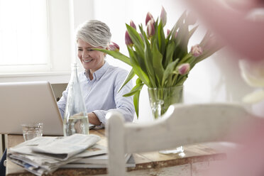 Reife Frau sitzt am Tisch und benutzt einen Laptop - FMKF001478