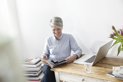 Ältere Frau sitzt am Tisch mit Zeitschrift und Laptop, lizenzfreies Stockfoto