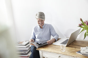 Ältere Frau sitzt am Tisch mit Zeitschrift und Laptop - FMKF001483