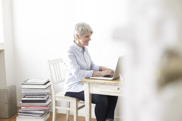 Reife Frau sitzt am Tisch und benutzt einen Laptop - FMKF001515