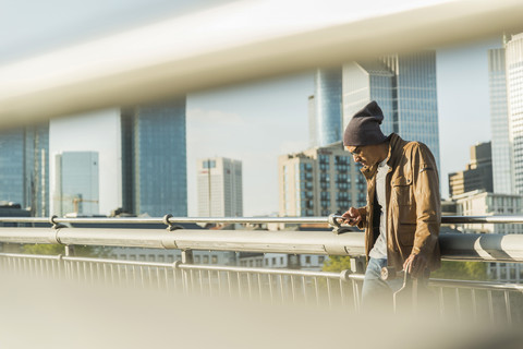 Deutschland, Frankfurt, Mann auf Brücke mit Skateboard schaut auf Smartphone, lizenzfreies Stockfoto