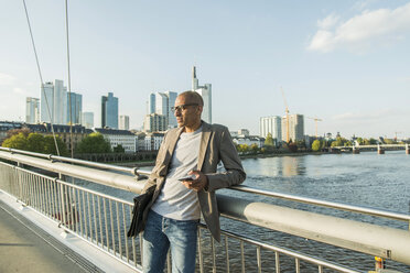 Deutschland, Frankfurt, Geschäftsmann auf Brücke mit Aktentasche und Smartphone - UUF004077