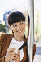 Porträt einer lächelnden Geschäftsfrau mit Smartphone, die an der Bushaltestelle wartet - EBSF000593
