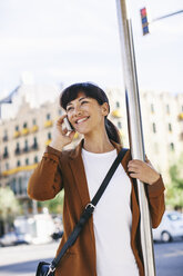 Spanien, Barcelona, Porträt einer lächelnden Geschäftsfrau, die an der Bushaltestelle mit ihrem Smartphone telefoniert - EBSF000595