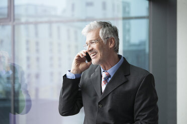 Businessman standing at window using mobile phone - RBF002665