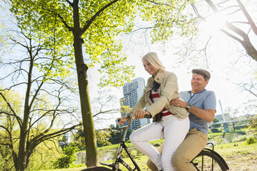 Mature couple riding bike in park, man sitting on rack - UUF004124