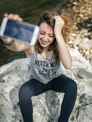 Porträt einer lachenden jungen Frau, die auf einem Felsen sitzt und ein Selfie mit einem Smartphone macht - MGOF000217