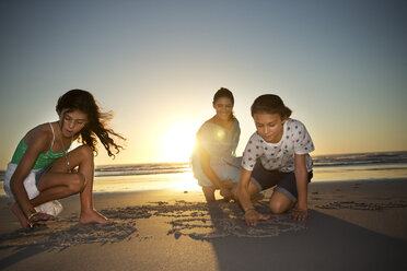 Mother with daughters on the beach at sunset drawing in sand - TOYF000280