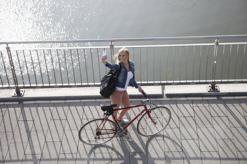 Deutschland, Düsseldorf, blonde Frau mit Fahrrad steht vor dem Rhein und macht ein Selfie mit Smartphone - RHF000815