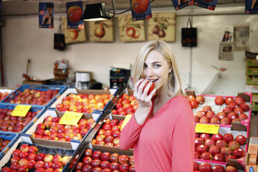 Lächelnde blonde Frau isst einen Apfel vor einem Obststand auf dem Wochenmarkt - RHF000822