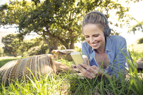 Lächelnde Frau liegt auf einer Wiese und hört Musik vom Smartphone, lizenzfreies Stockfoto