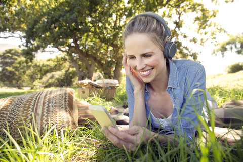Lächelnde Frau liegt auf einer Wiese und hört Musik vom Smartphone, lizenzfreies Stockfoto