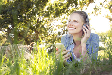 Lächelnde Frau liegt auf einer Wiese und hört Musik vom Smartphone - TOYF000369