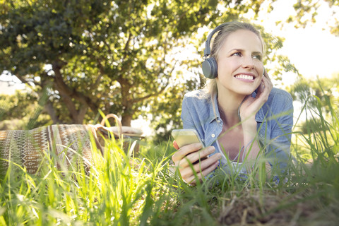 Lächelnde Frau liegt auf einer Wiese und hört Musik vom Smartphone, lizenzfreies Stockfoto