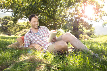 Smiling man lying on meadow reading book - TOYF000393