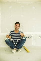 Smiling young man sitting at blank wall holding pocket rule - UUF004184