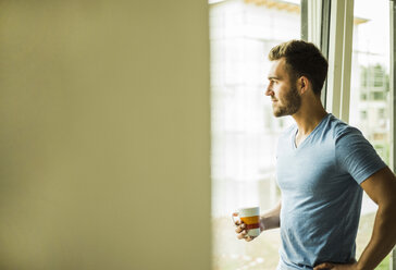 Junger Mann hält Kaffeetasse und schaut aus dem Fenster - UUF004239