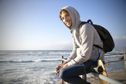 Smiling young man sitting on railing at the coast - TOYF000440