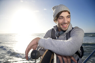 Lächelnder junger Mann mit Longboard an der Küste - TOYF000442
