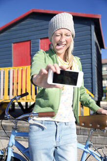 Verspielte junge Frau macht ein Selfie mit Fahrrad vor einer bunten Strandhütte - TOYF000459