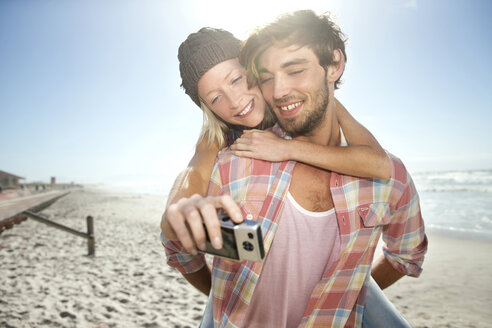 Junge Frau auf dem Rücken ihres Freundes am Strand macht ein Selfie - TOYF000487