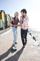 Junger Mann hilft seiner Freundin auf dem Skateboard auf der Strandpromenade - TOYF000502