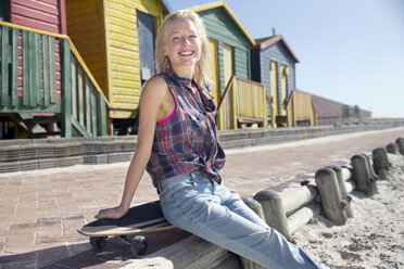 Glückliche junge Frau mit Skateboard auf der Strandpromenade sitzend - TOYF000504