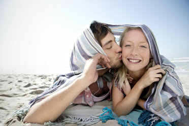 Glückliches junges Paar liegt am Strand und küsst sich unter einer Decke - TOYF000508